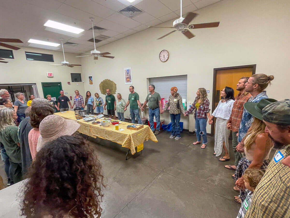 A group of people stand in a circle around tables covered with yellow tablecloths and food trays in a brightly lit room, preparing for a communal meal—a warm gathering organized by Kokua Tree Care to celebrate tree care Maui initiatives.