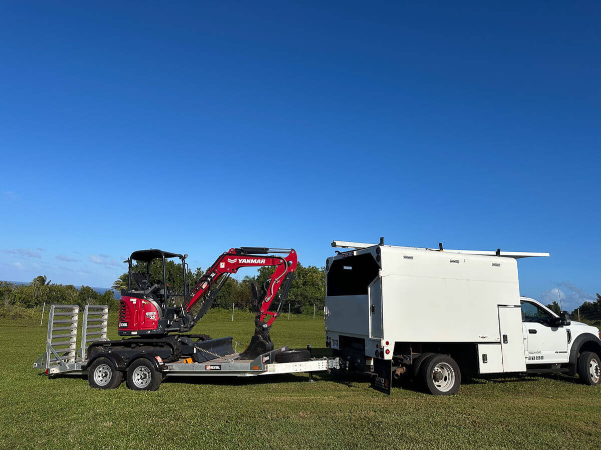A white utility truck is parked on grass, towing a trailer with a small red and black Yanmar excavator—perfect for kokua tree care or tree care Maui. The sky is clear and blue, with green trees and fields in the background.
