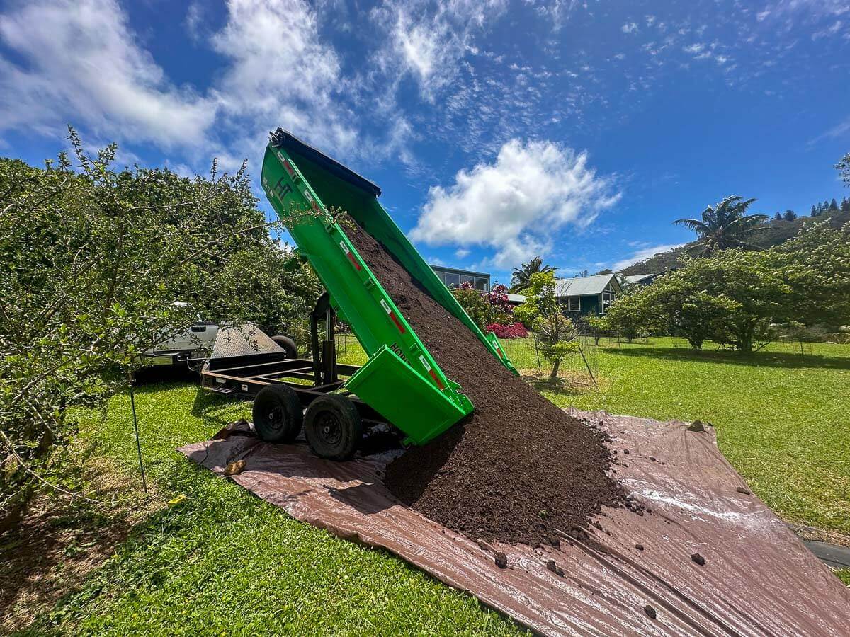 A green dump trailer from Kokua Tree Care unloads a large pile of soil onto a brown tarp on a grassy lawn, with trees and a house in the background under a bright, partly cloudy sky—perfect for expert tree care Maui services.